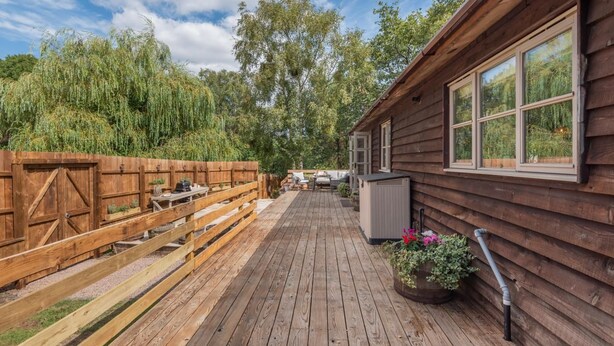 Een houten terras met buitenmeubilair, een gebouw met grote ramen en een natuurlijke achtergrond.