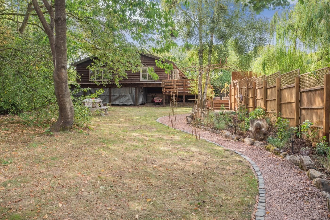 Eine Holzhütte mit einer Laube, umgeben von einem Garten mit Kiesweg und Holzzaun.