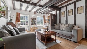 Ein gemütliches Wohnzimmer mit sichtbaren Holzbalken, einer Couch, einem Sessel, einem Couchtisch und gerahmten Bildern an den Wänden.