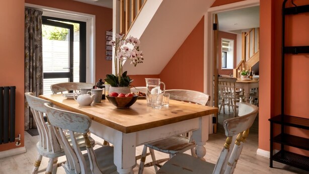 Un coin repas avec une table en bois, des chaises blanches, un vase de fleurs et une vue sur un escalier.