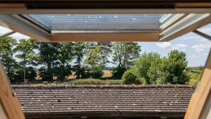 Vue depuis une lucarne d’une pièce où se trouvent un banc en bois et une petite table, donnant sur un jardin avec des arbres et une clôture.