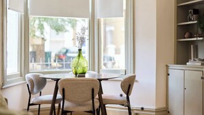 Een eetgedeelte met een ronde tafel, twee stoelen, een groene vaas met gedroogde bloemen en uitzicht door grote ramen.