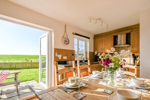 A dining area with a table set for two, wooden chairs, a bench, and a view of a green field through a glass door. - 3 Bed in Pen Y Cwm (oc-d34208) (Newgale)