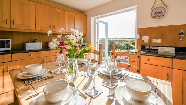 Un coin repas avec une table dressée pour quatre personnes, un vase de fleurs, des meubles en bois et une vue à travers une porte vitrée.