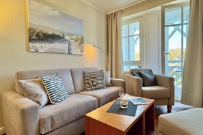 A living room with a sofa, armchair, and coffee table, a painting on the wall, and a window with curtains. - Ferienwohnung mit Fitness und Freizeitangeboten (Sellin)