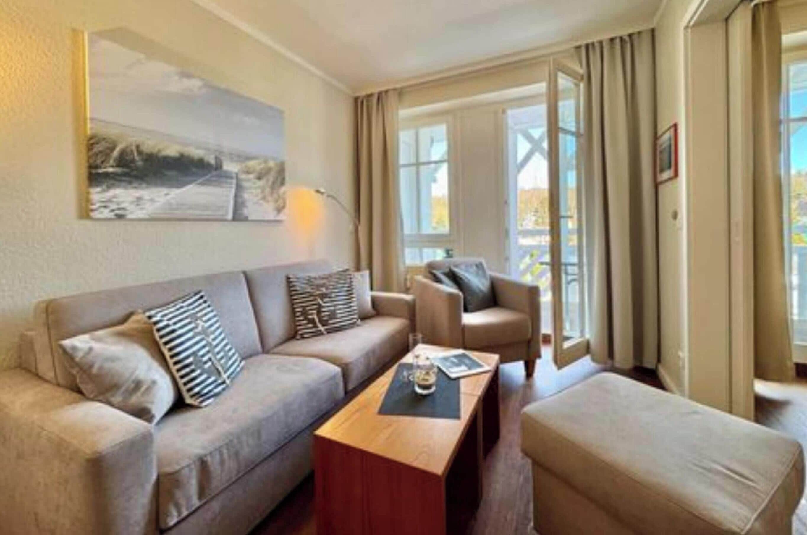 Et værelse med sofa, lænestol, skammel og sofabord. Et maleri hænger på væggen, og der er et vindue med gardiner.
