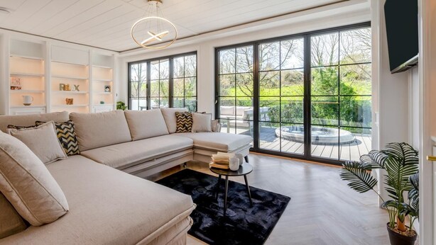 A modern living room with a sectional sofa, a coffee table, and a large window with a view of a garden and hot tub. - 2 Bed in St. Minver (oc-g34708) (Saint Minver)