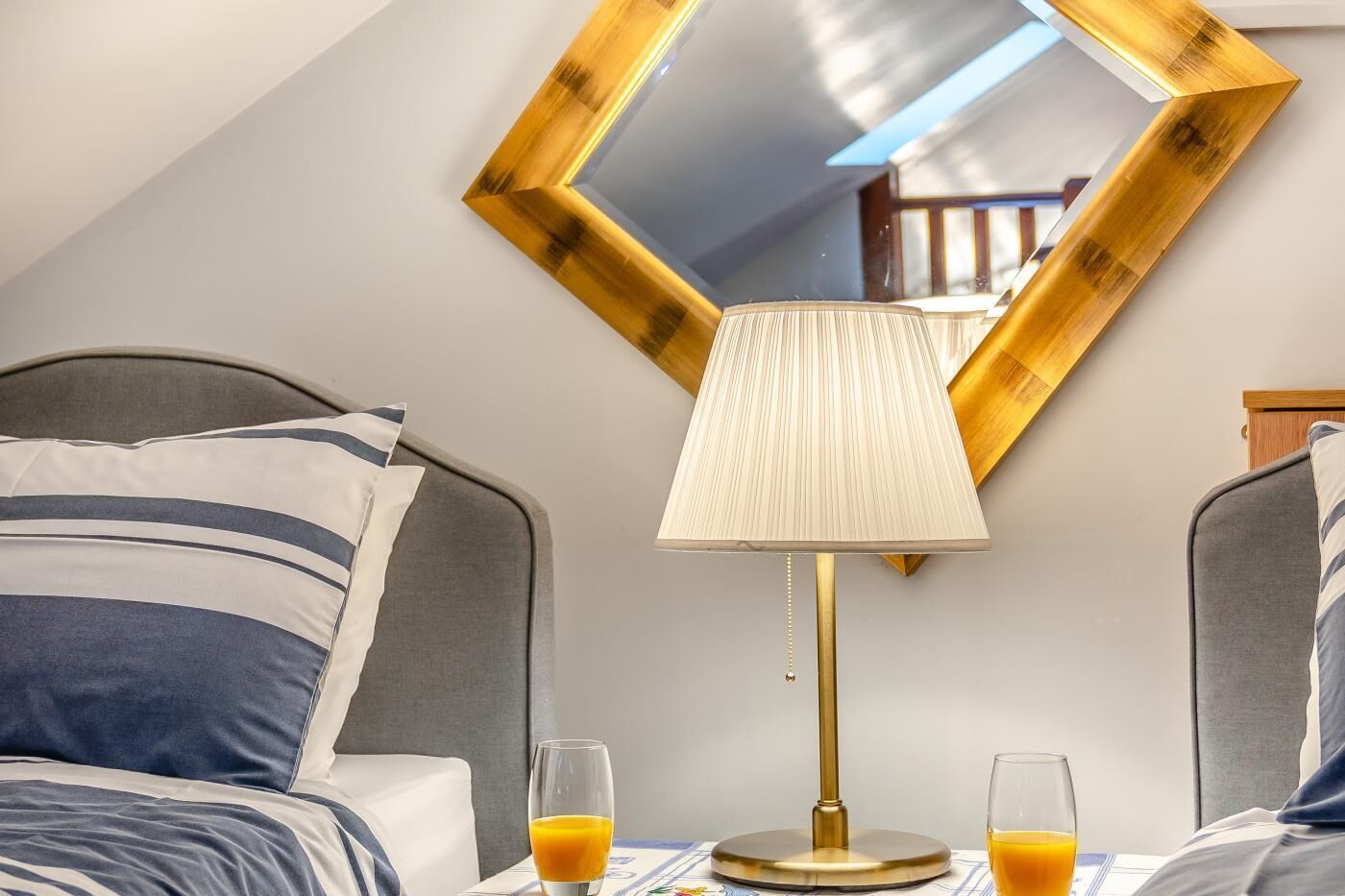 A hotel room with a bed, a bedside table with a lamp, two glasses of orange juice, and a large mirror on the wall.