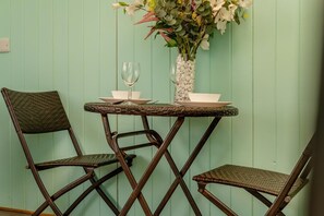 A room with a light green wall, a small round table with a glass vase and a wine glass, and two wicker chairs. - 1 Bed in Bude (oc-a34844) (Bude)