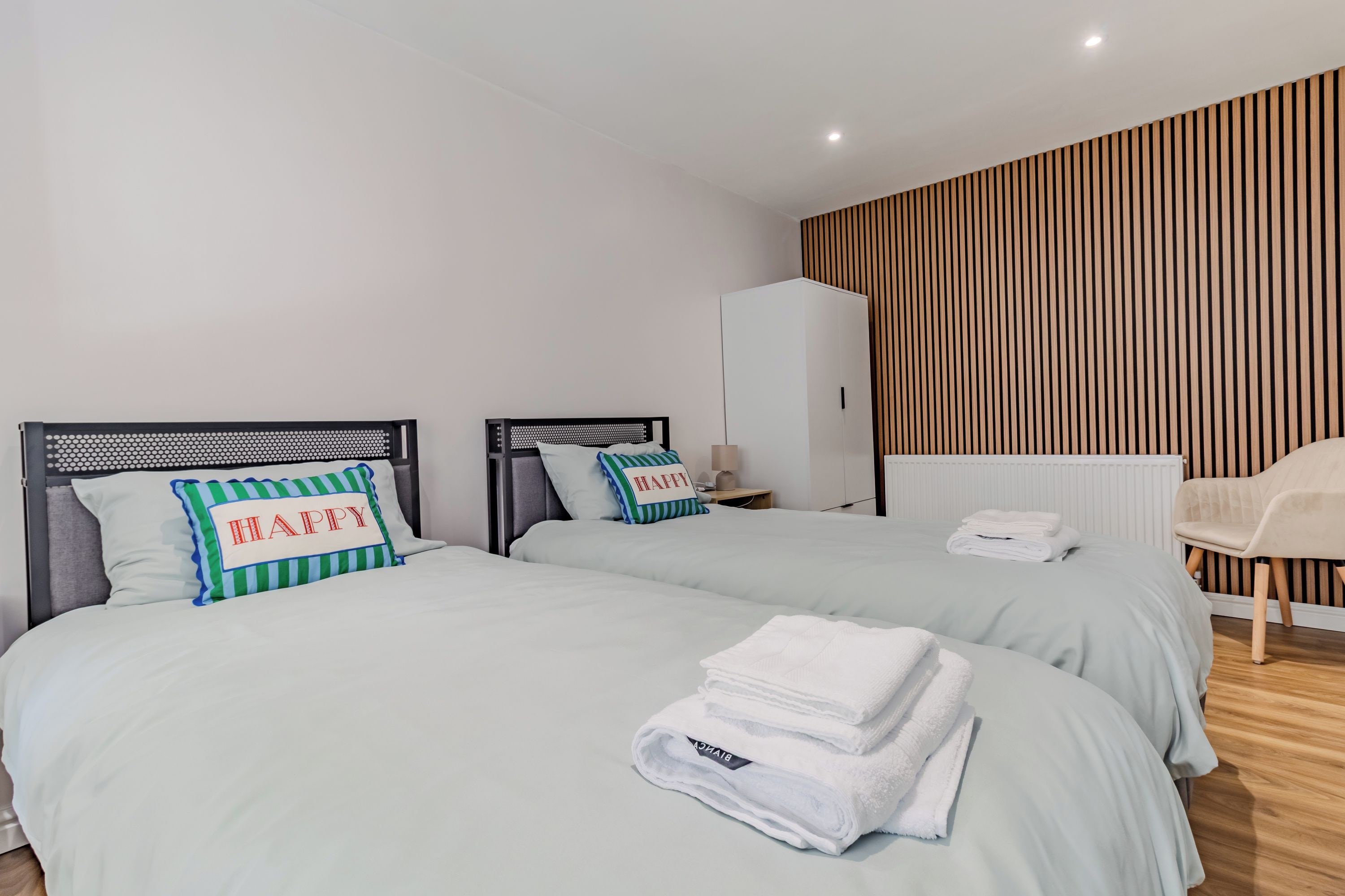 A hotel room with two beds, a wardrobe, a chair, and a radiator.