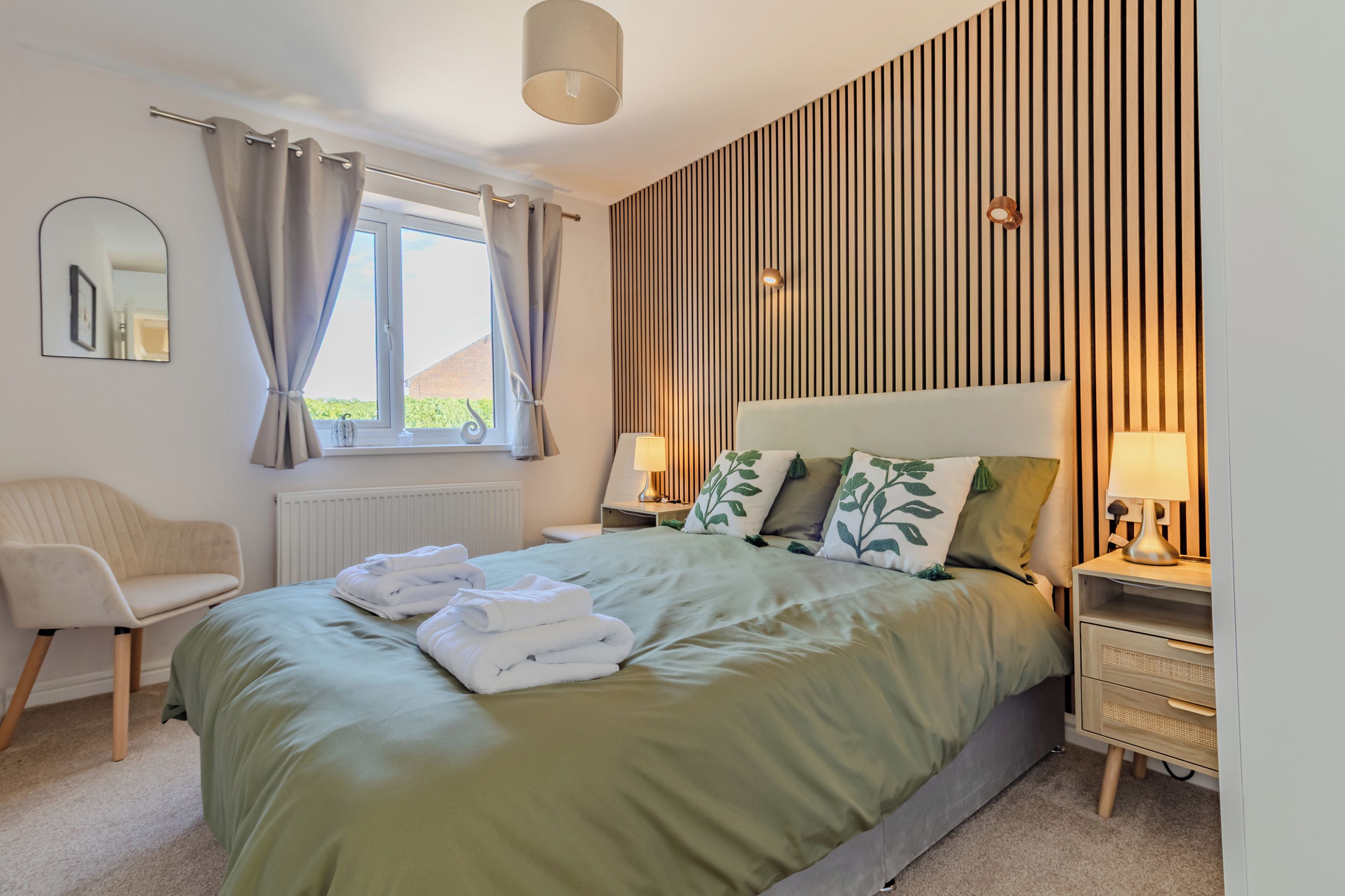 A bedroom with a bed, bedside tables, a chair, a window with curtains, and a wall with vertical wooden slats.