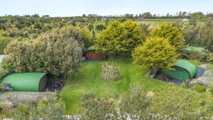 A rural setting with multiple green-roofed structures, likely cabins, surrounded by lush greenery and a clear sky. - 1 Bed in Bude (oc-s34854) (Bude)