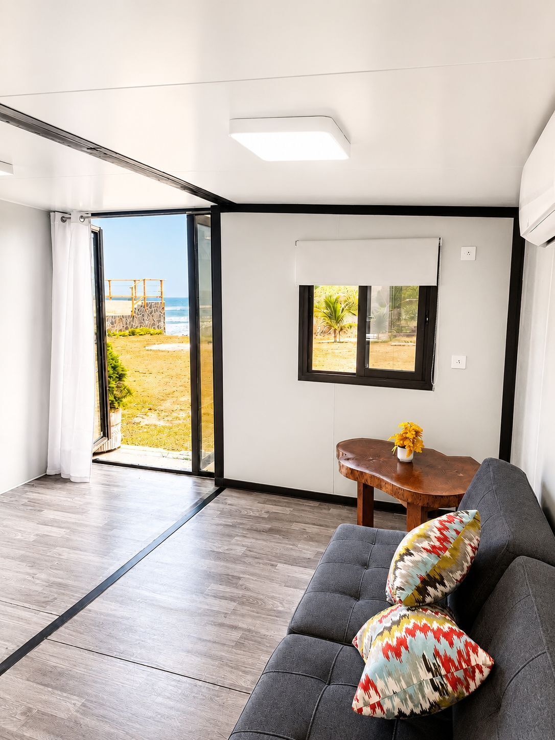 A modern interior with a grey sofa, a wooden table with a vase, and a view of a beach through large glass doors.