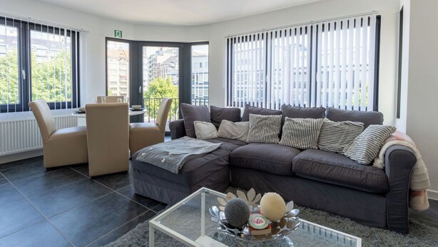 Ein modernes Wohnzimmer mit einem Ecksofa, einem Glastisch und Blick auf die Stadt durch große Fenster.