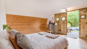 Ein Schlafzimmer mit Holz-Kopfteil, einem Bett mit weißer Bettwäsche, einem kleinen Schreibtisch mit Computer und Blick auf einen Garten durch eine Tür.