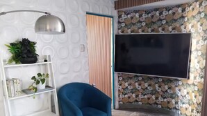 A living room with a blue armchair, a white shelving unit, a flat-screen TV, and a floral wallpaper. - 4. Moderno Departamento Entero Estilo Minimalista - CUARTO PISO (Breña)
