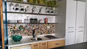 A kitchen with a tiled backsplash, stainless steel sink, and a gas stove. There are shelves above the sink with various kitchen items and a green bowl on the counter. - 4. Moderno Departamento Entero Estilo Minimalista - CUARTO PISO (Breña)