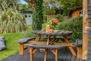 Een houten buitentafel met banken, omgeven door groen en een houten constructie op de achtergrond.