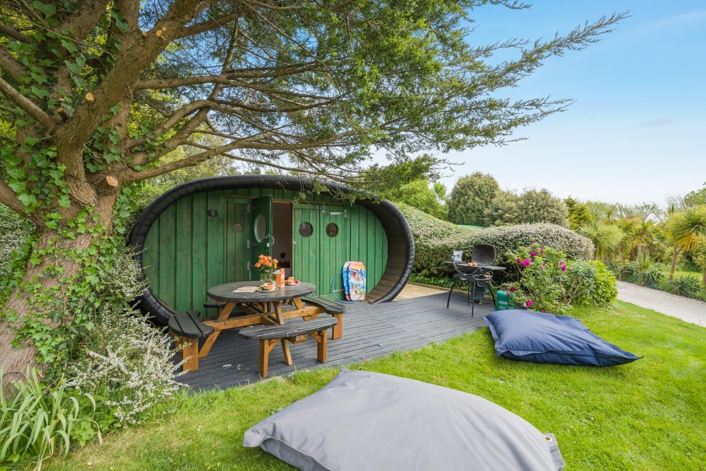 Una estructura de madera verde con una puerta circular, una mesa de picnic y una parrilla de barbacoa en el exterior.