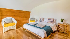 Ein Schlafzimmer mit Holzboden, einem Bett mit blauer Decke, einem Korbstuhl mit gelber Sitzkissen und einem kleinen Holfnachtkästchen mit einer Pflanze.