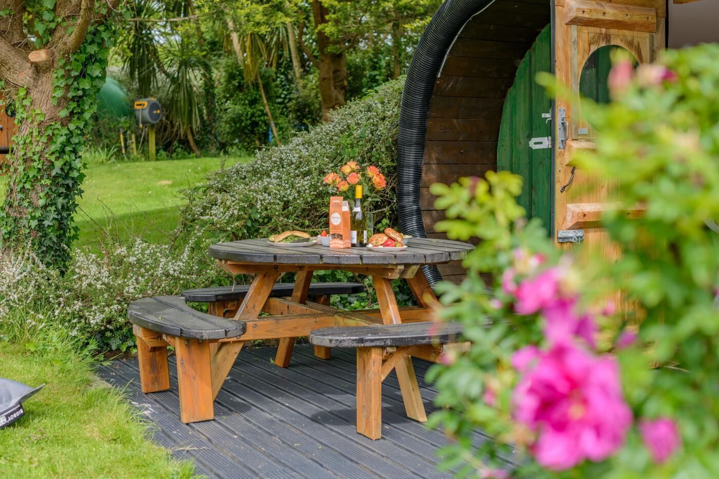 Een houten picknicktafel met bankjes, omgeven door groen en een groene deur.
