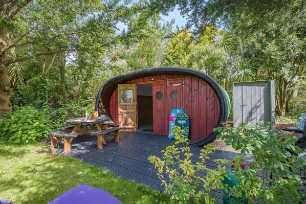 A small, round wooden structure with a door, a picnic table, and a surfboard outside.