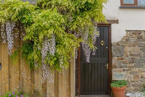 Una cerca de madera con follaje verde y flores moradas colgantes, una puerta negra con un aldabón de latón y un muro de piedra con una planta en maceta.