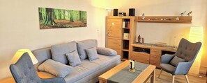 Ein Wohnzimmer mit einer Couch, einem Sessel, einem Couchtisch, einem Bücherregal, einer Lampe und einem Waldgemälde.