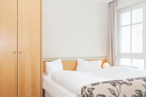 A hotel room with a bed, a wooden wardrobe, a window with curtains, and a bedside table with a lamp. - Ferienwohnung mit Moblierten Balkon in Sellin (Sellin)