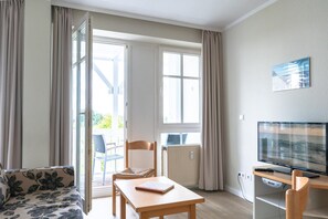 A room with a sofa, a table with a book, a chair, a TV, and a window with curtains. - Ferienwohnung mit Moblierten Balkon in Sellin (Sellin)
