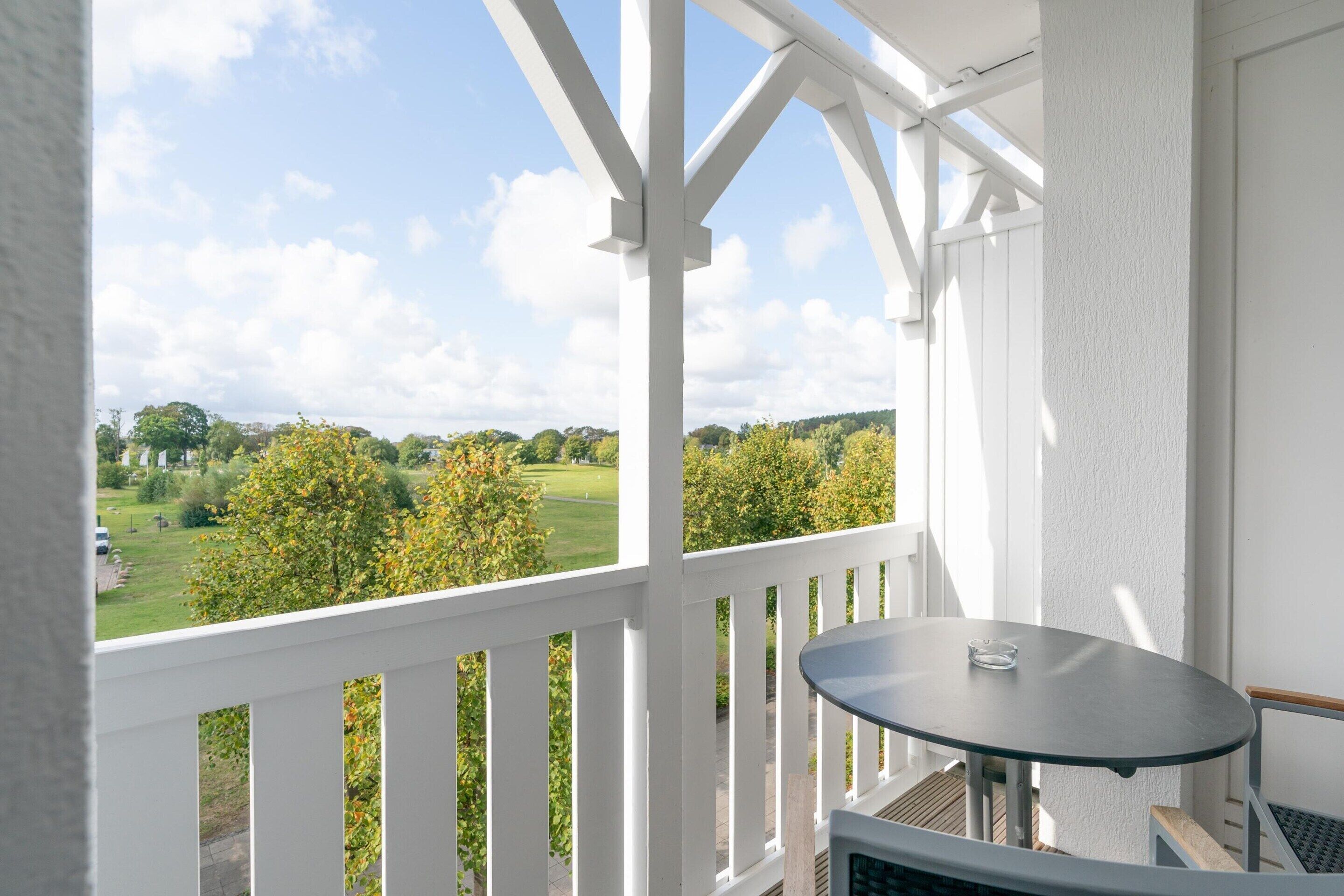 A balcony with a table and chairs, overlooking a green landscape with trees and a clear sky.