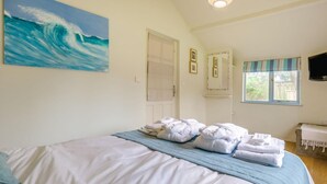 Quarto com cama, toalhas, pintura de uma onda, janela com cortinas e televisão.