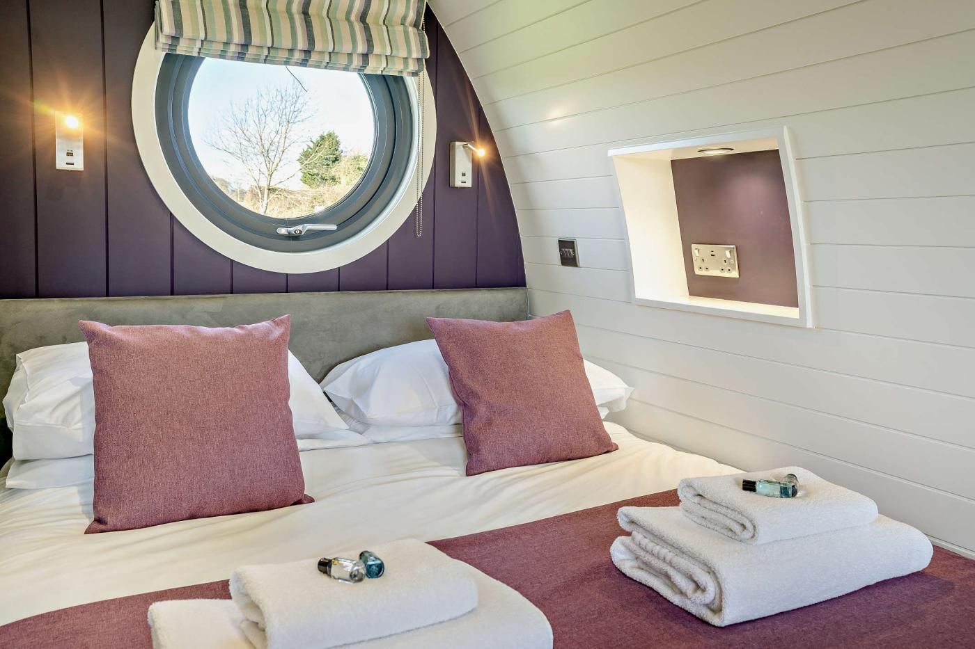 A bedroom with a round window, a bed with white and pink pillows, a bedside table with a lamp, and a view of the outdoors.