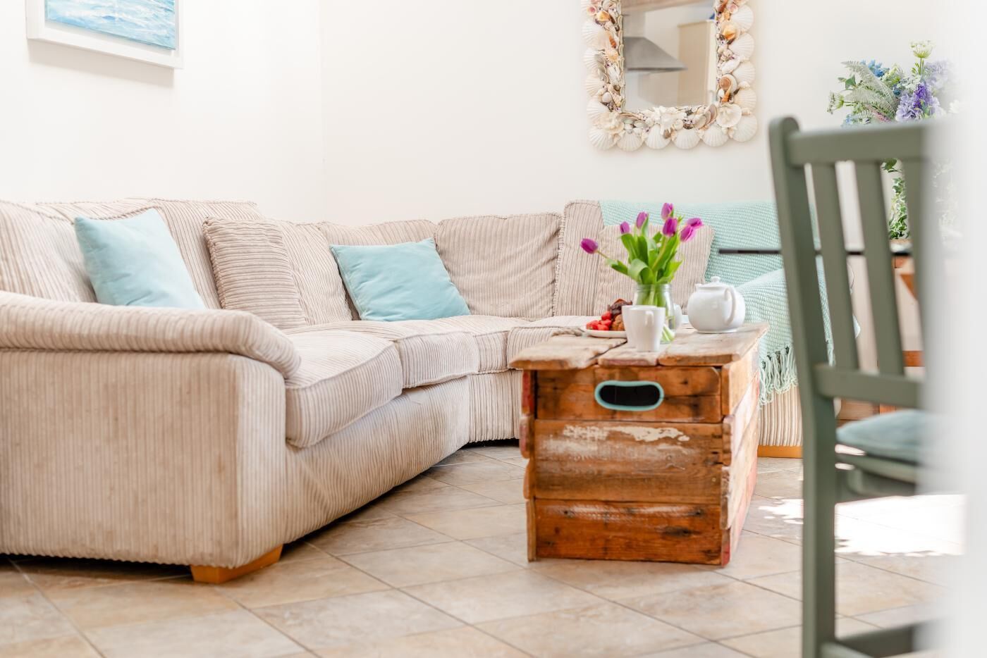 Ein beiges Sofa mit Cordstoff, zwei hellblaue Kissen, eine hölzerne Kommode, ein weißer Teekessel und eine Vase mit violetten Blumen.