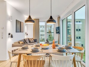 A modern dining area with a large table, chairs, and pendant lights. There is a sofa with cushions and a TV mounted on the wall. - Cosy apartment for 6 guests with WIFI, TV, balcony and pets allowed (Diepholz)