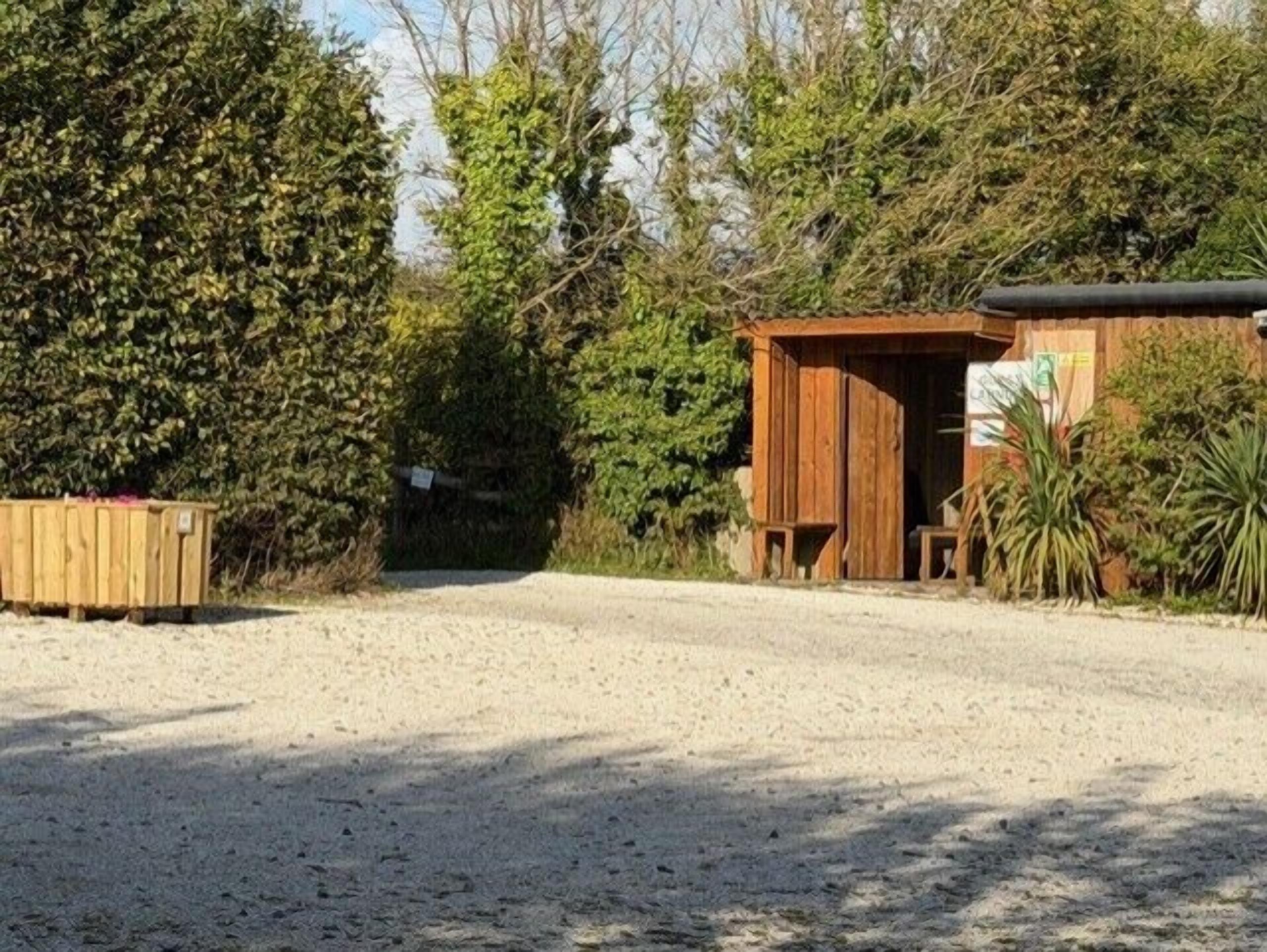 Ein Schotterweg führt zu einer Holzhütte mit einer Tür, umgeben von dichtem Grün.