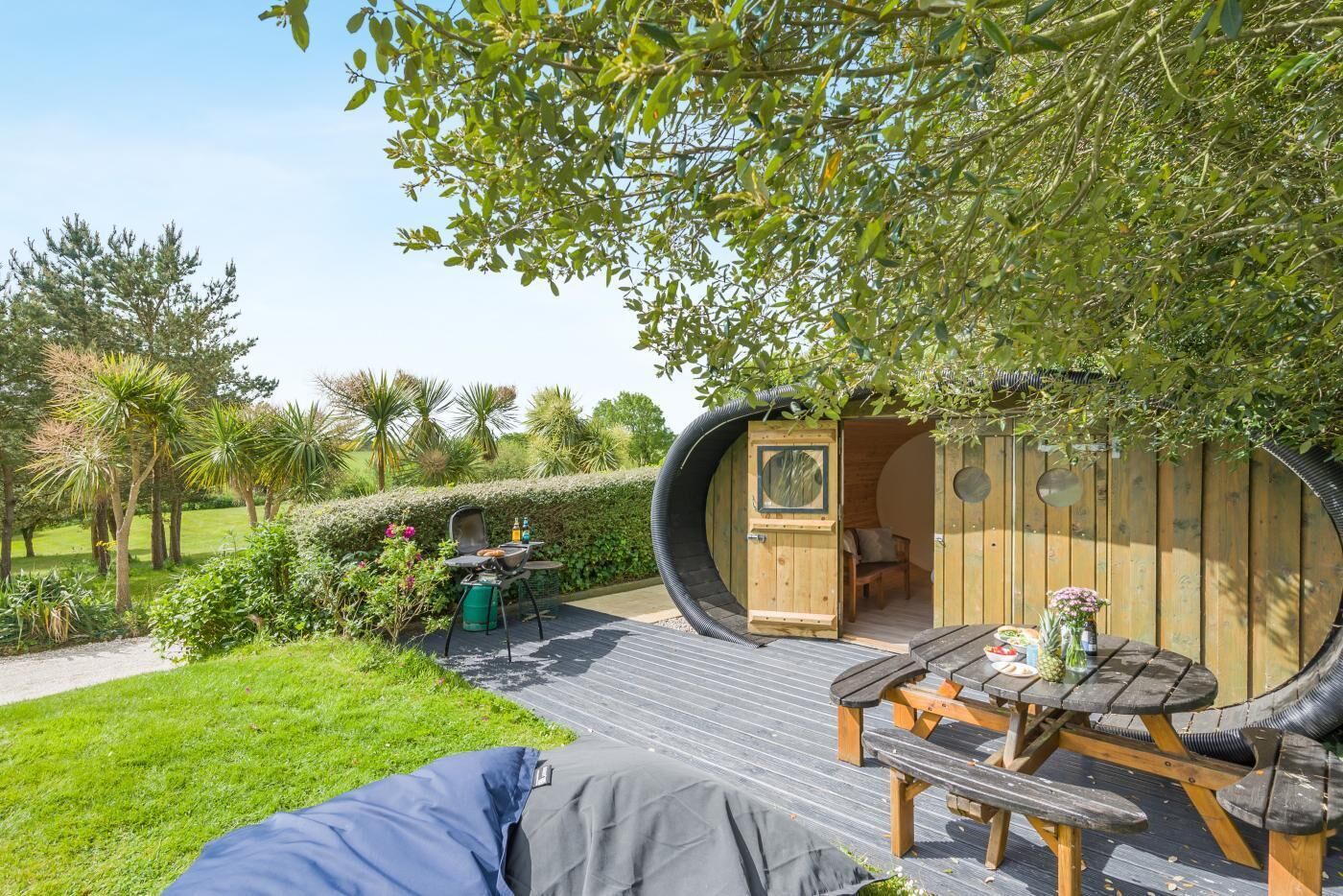 A circular wooden structure with a bench and table, surrounded by a garden with various plants and a lawn.