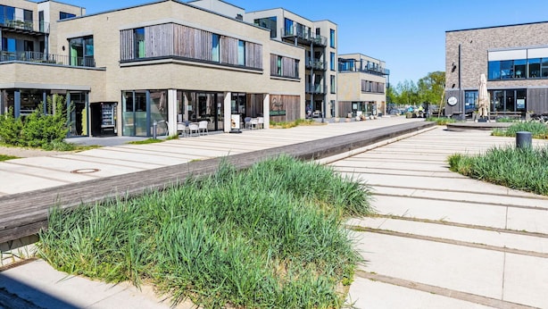 Eine moderne Stadtstraße mit mehrstöckigen Gebäuden, Außenbestuhlung und blauem Himmel.