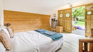 Ein Schlafzimmer mit einem Holz-Kopfteil, einem Bett mit weißen Bettwäsche, einem kleinen Holzschemel und Blick auf einen Außenbereich durch eine Holztür.