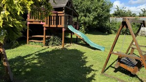 Ein hölzernes Spielhäuschen mit Rutsche, Schaukel und einem Baum im Vordergrund.