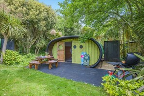 Eine kleine, grüne, kuppelartige Struktur mit einer Tür, umgeben von einem Garten mit Picknicktisch und Grill.