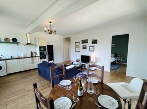 A modern kitchen with white cabinets, a microwave, and a sink. There is a dining area with a wooden table set for four, a sofa, and a TV.