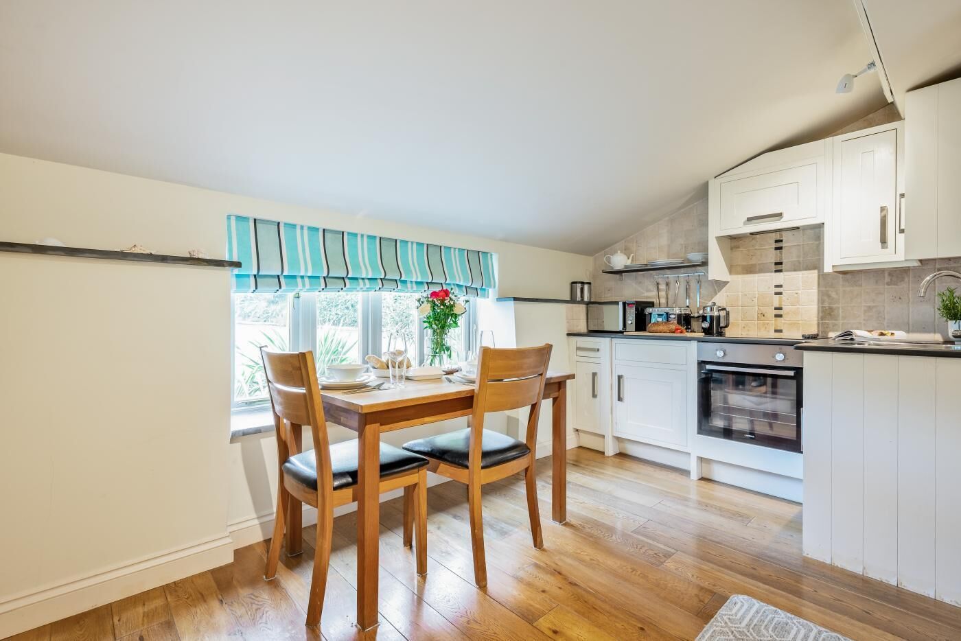 Een kleine keuken met houten kastjes, een eettafel met stoelen en een raam met gordijnen.