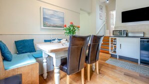 A dining area with a table, chairs, a painting, and a kitchen with appliances. - 2 Bed in Bude (oc-c34841) (Bude)