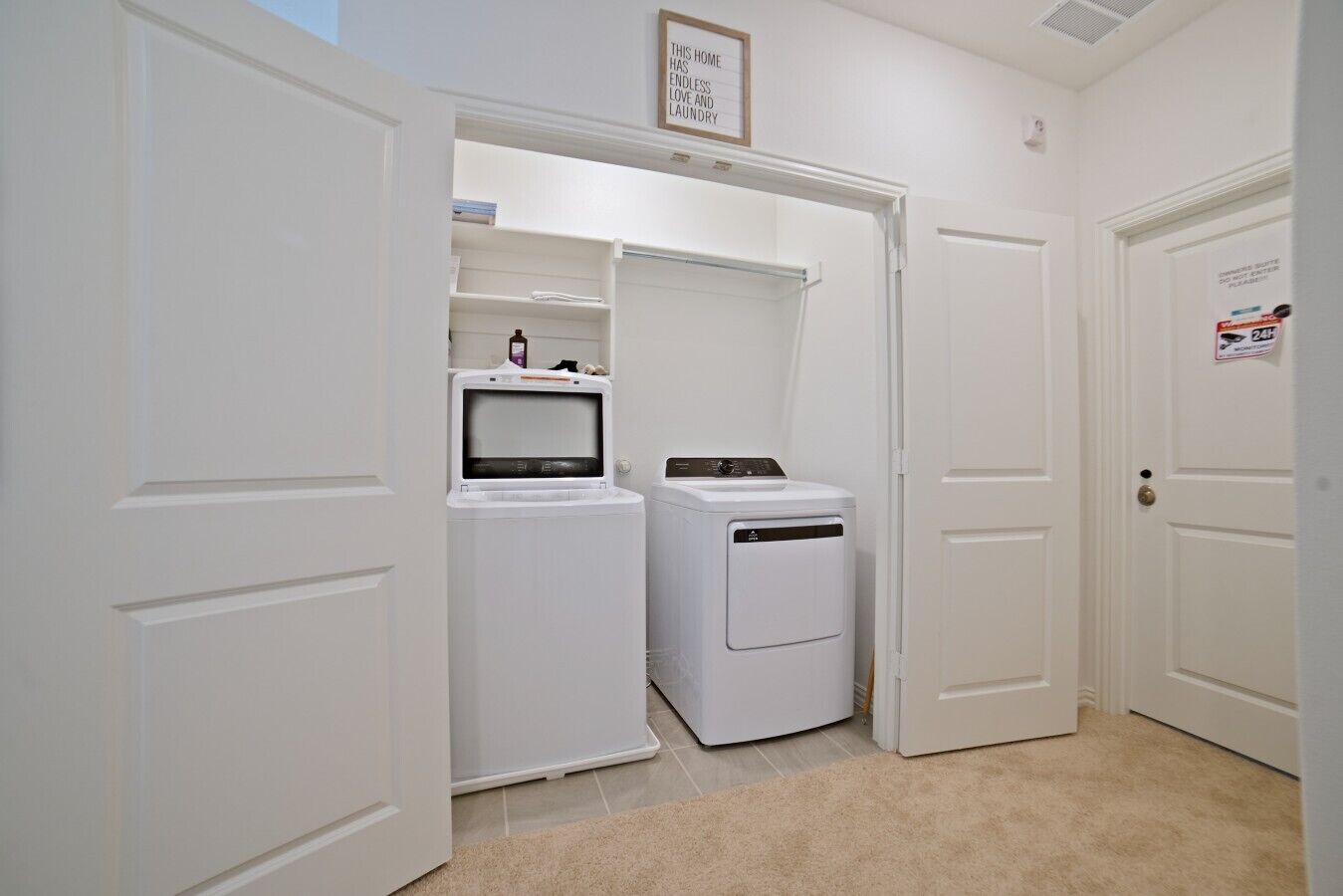 A laundry room with a washer and dryer, a shelf with laundry supplies, and a door with a sign.