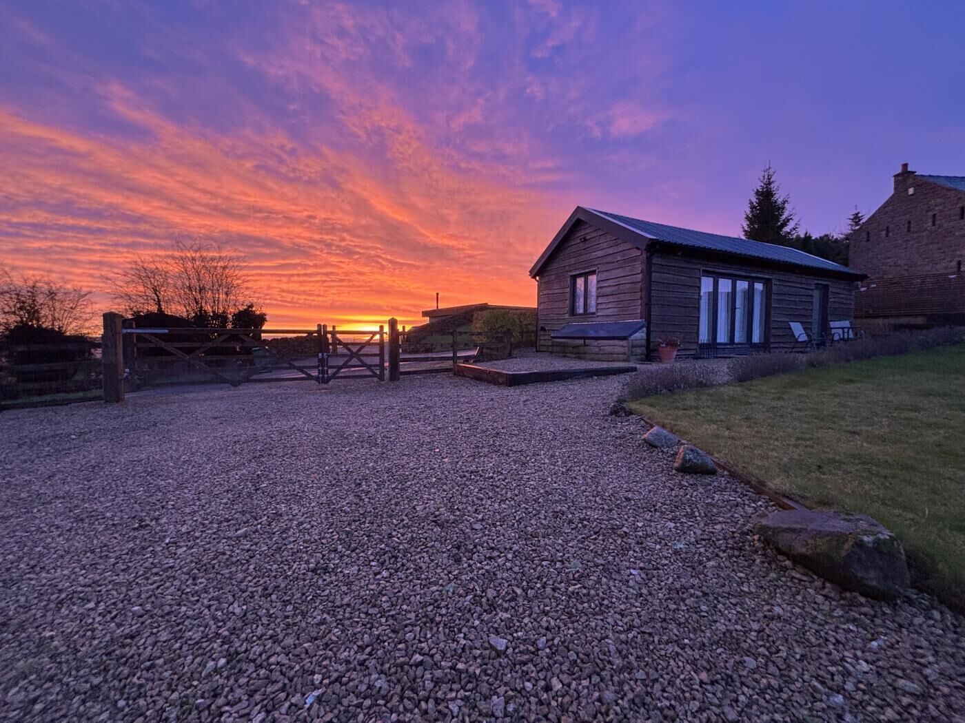 A gravel driveway leading to a wooden house with a metal gate, surrounded by a grassy area and a stone wall.