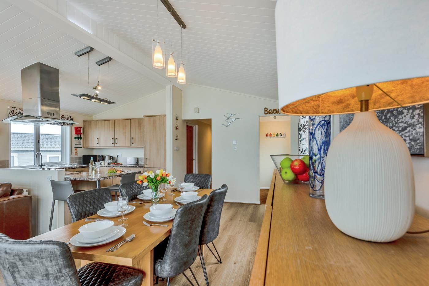 Una cocina moderna con armarios de madera, un comedor con una mesa de madera dispuesta para cuatro personas, y un jarrón grande con flores.