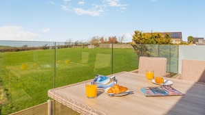 Un balcon avec une table préparée pour le petit-déjeuner, comprenant des croissants et du jus d’orange, donnant sur un champ verdoyant et un bâtiment situé au loin.