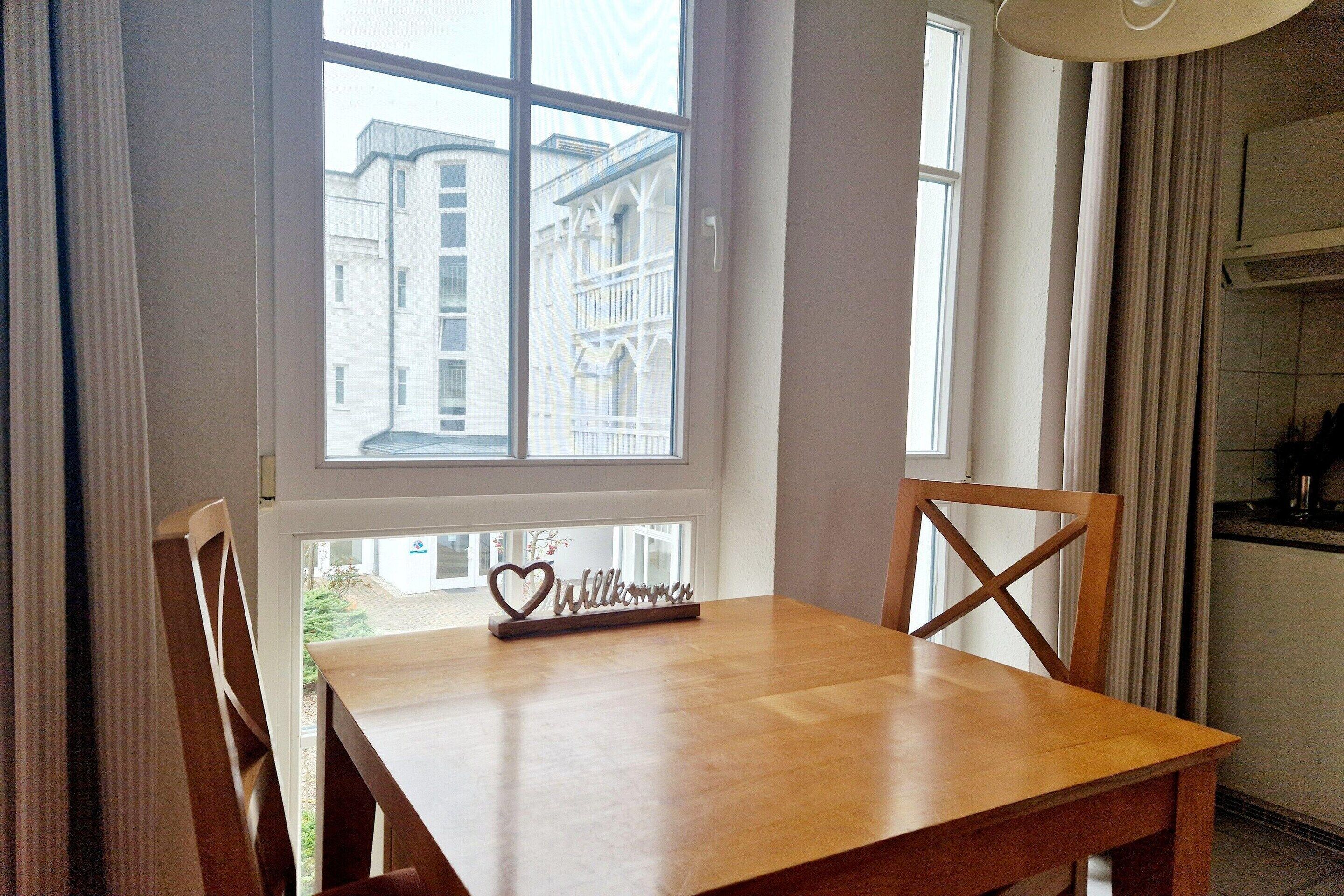 A wooden dining table with a heart-shaped welcome sign, two wooden chairs, and a window with a view of buildings.