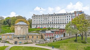 Un bâtiment historique doté d’une tour centrale, d’une allée pavée et d’un espace verdoyant avec des arbres.